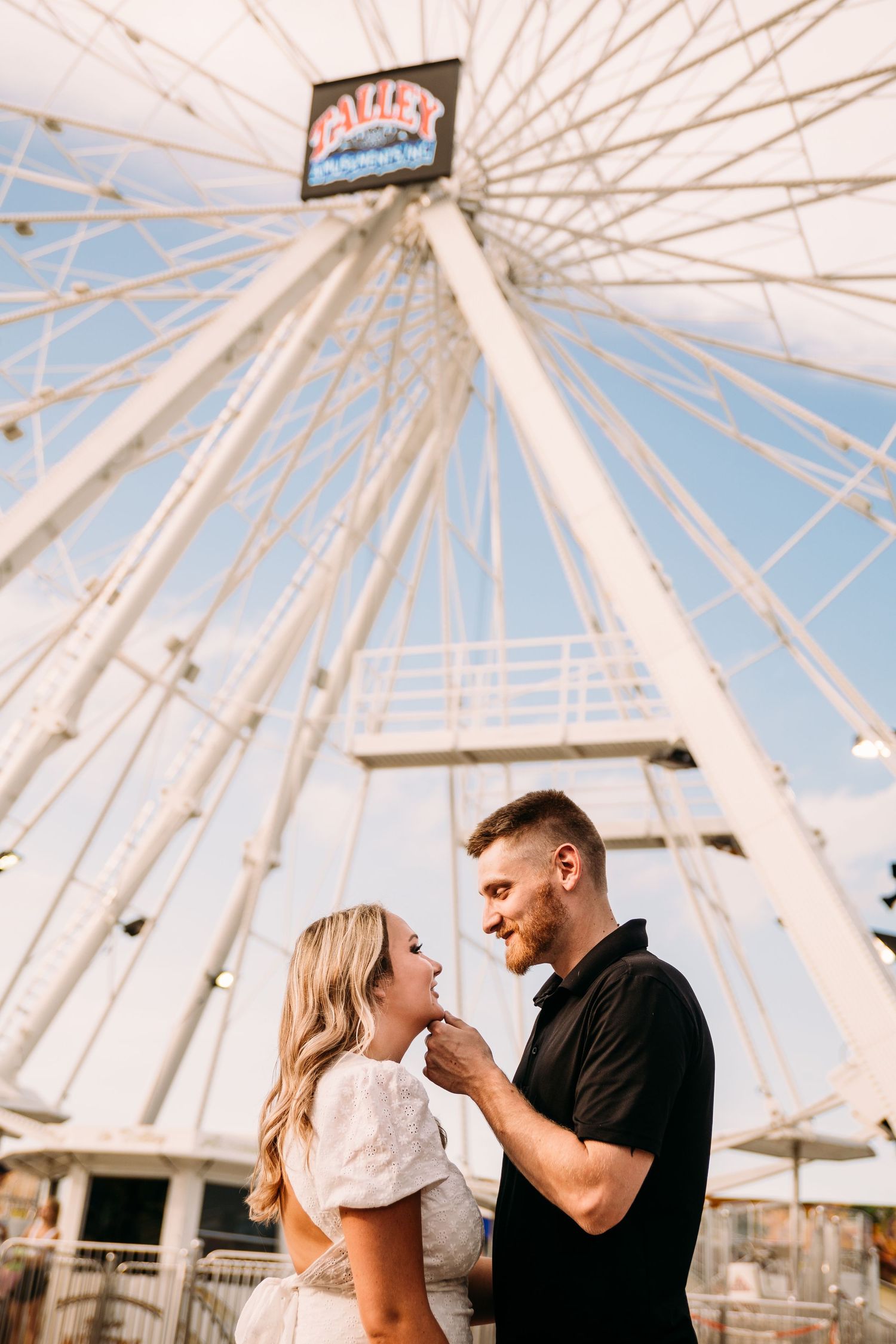 Romantic couple sharing an intimate moment beneath a large white Ferris wheel against a blue sky.