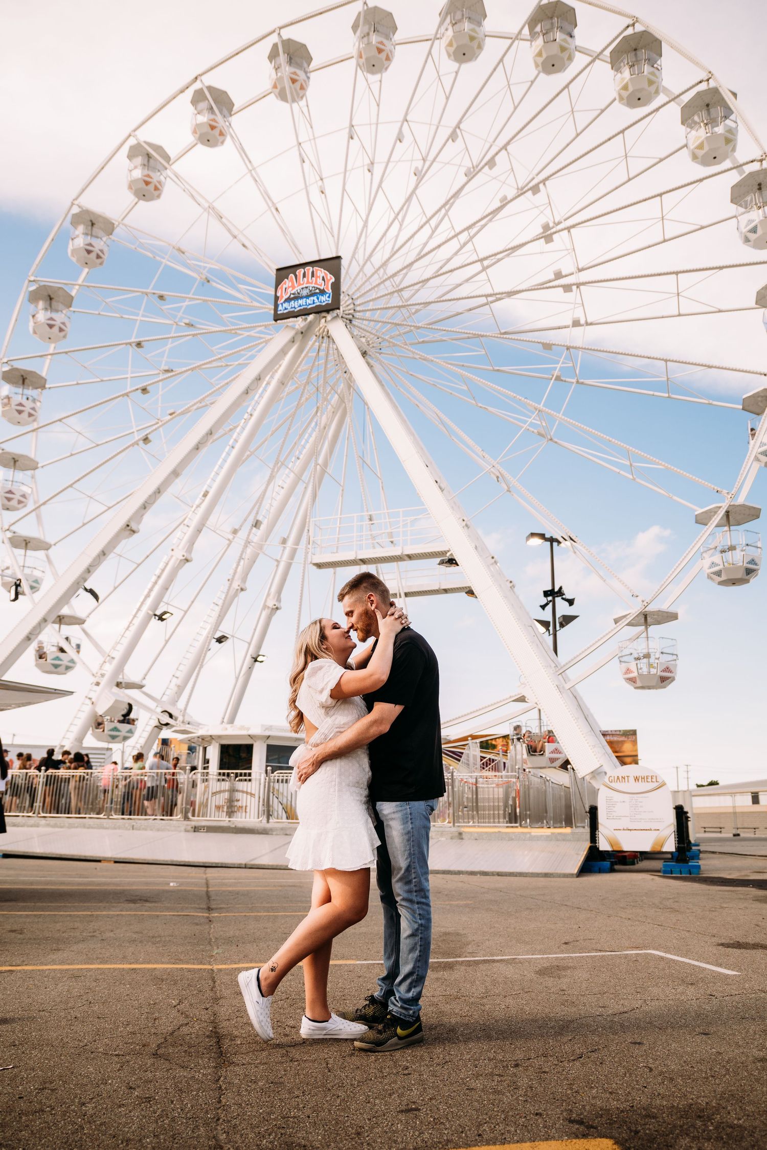 A couple embraces in front of a large white ferris wheel at a fairground.