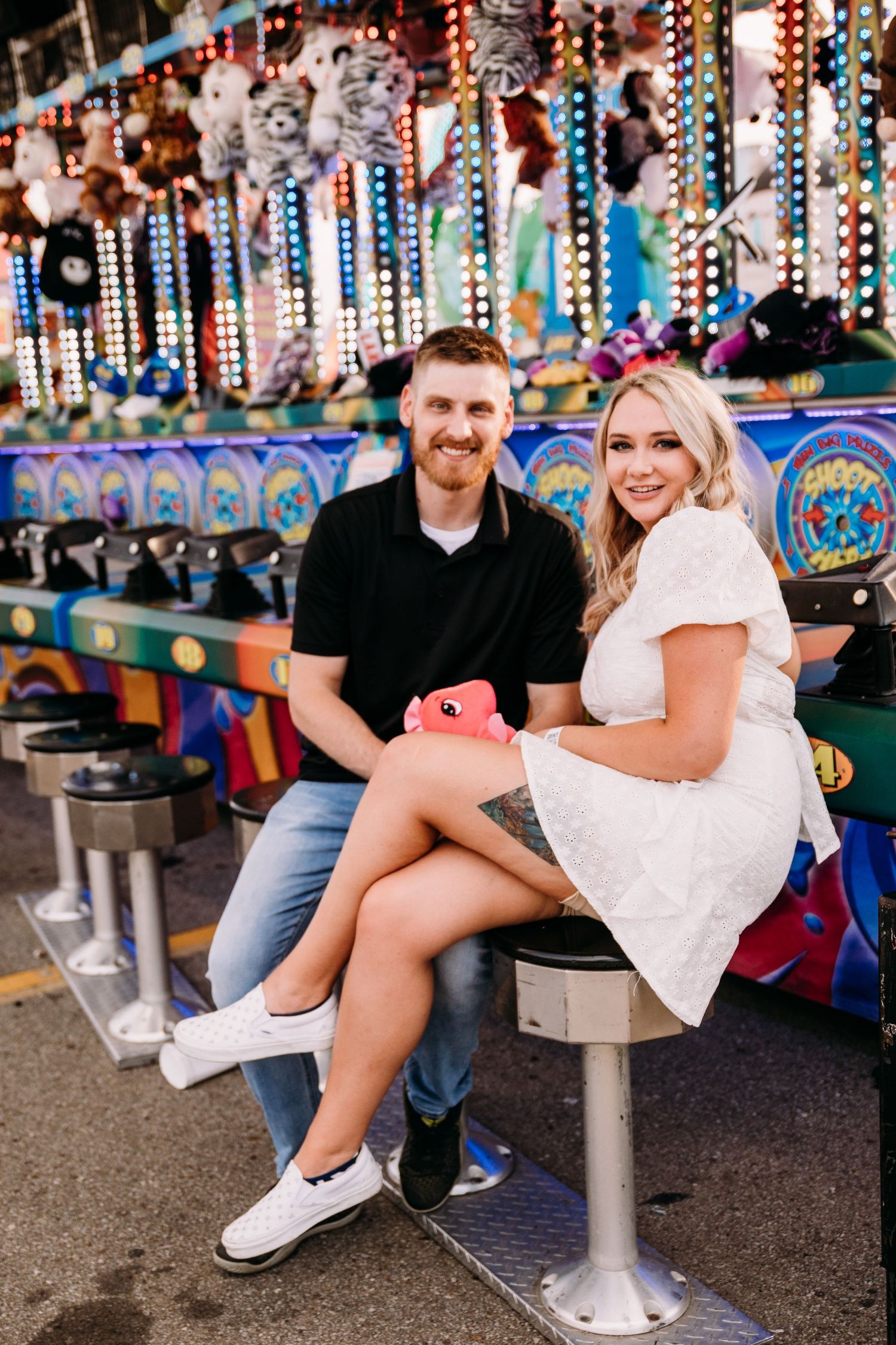 A couple sits together on stools at a colorful carnival game booth.