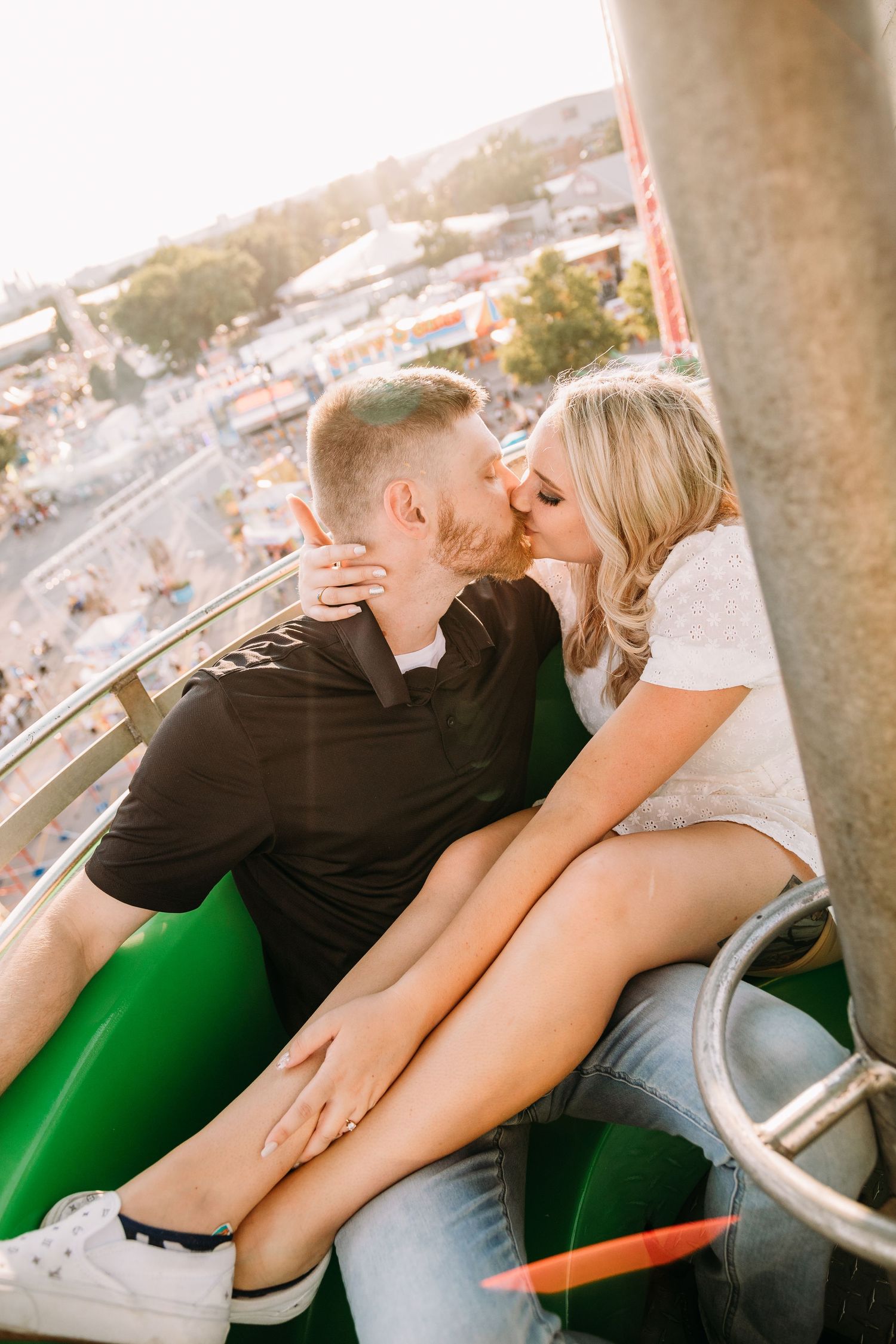 Couple sharing a tender kiss while sitting on a green carnival ride with city views below.