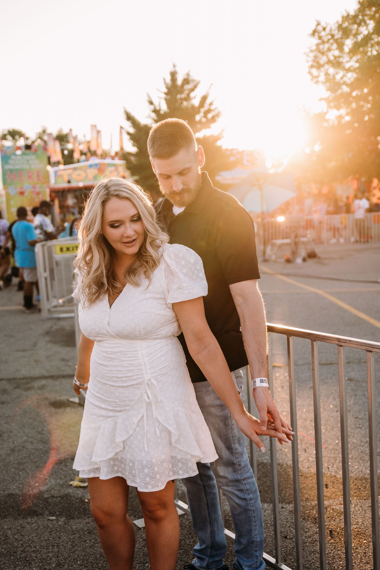 A couple embraces at sunset near carnival barriers with rides in background.