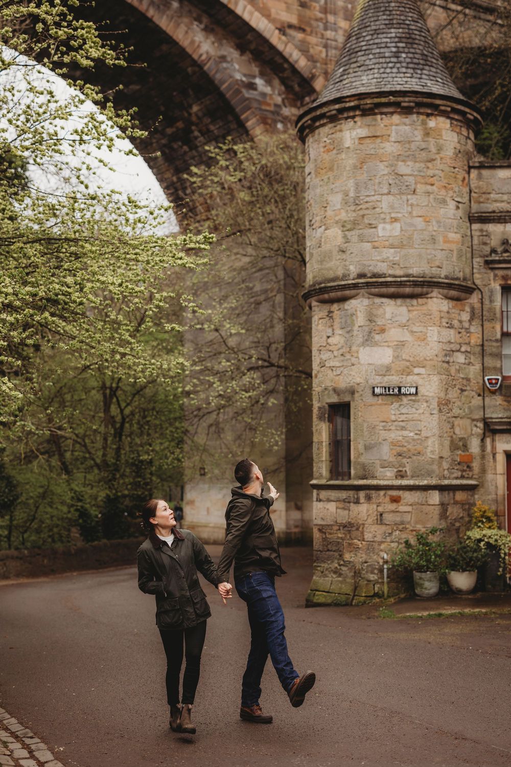Two people walking along a path beside a stone turret and archway of a historic building in warm autumn lighting.