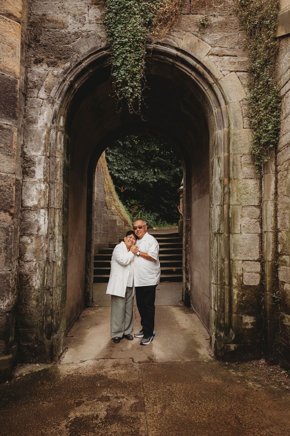 A pair in white clothing stands embracing under an ivy-covered stone archway.