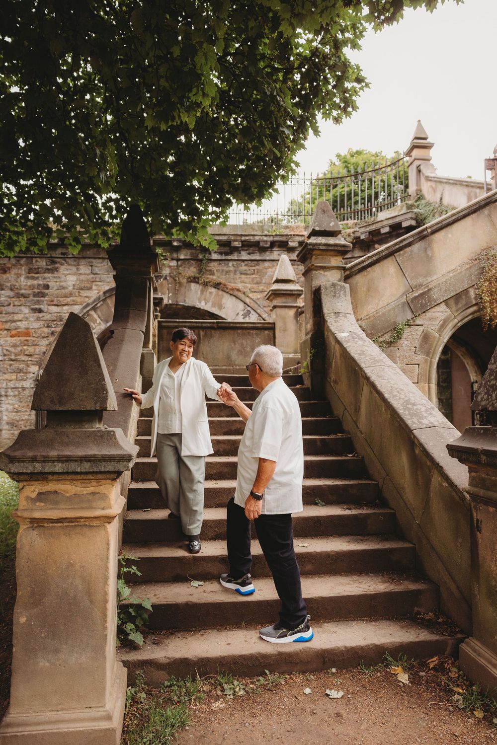 Two people in casual attire stand on historic stone steps with arched architectural details and trees in the background.