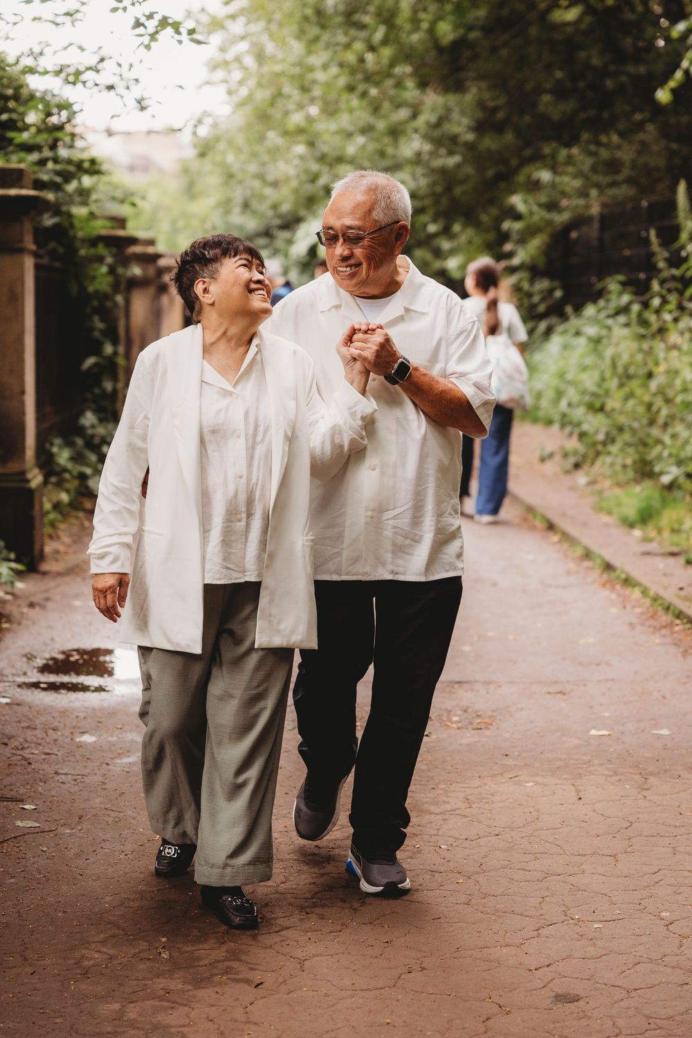 Senior couple in white outfits walking together and laughing on a garden path among trees.
