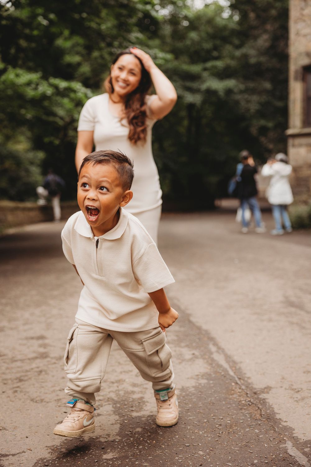 An energetic young child in beige outfit makes an excited expression while playing outdoors on a path with others visible behind.