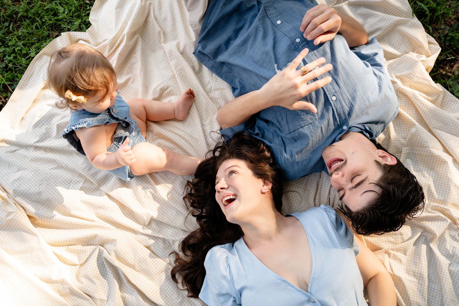 A family laughs together while laying on a blanket outdoors enjoying quality time together on a sunny day.