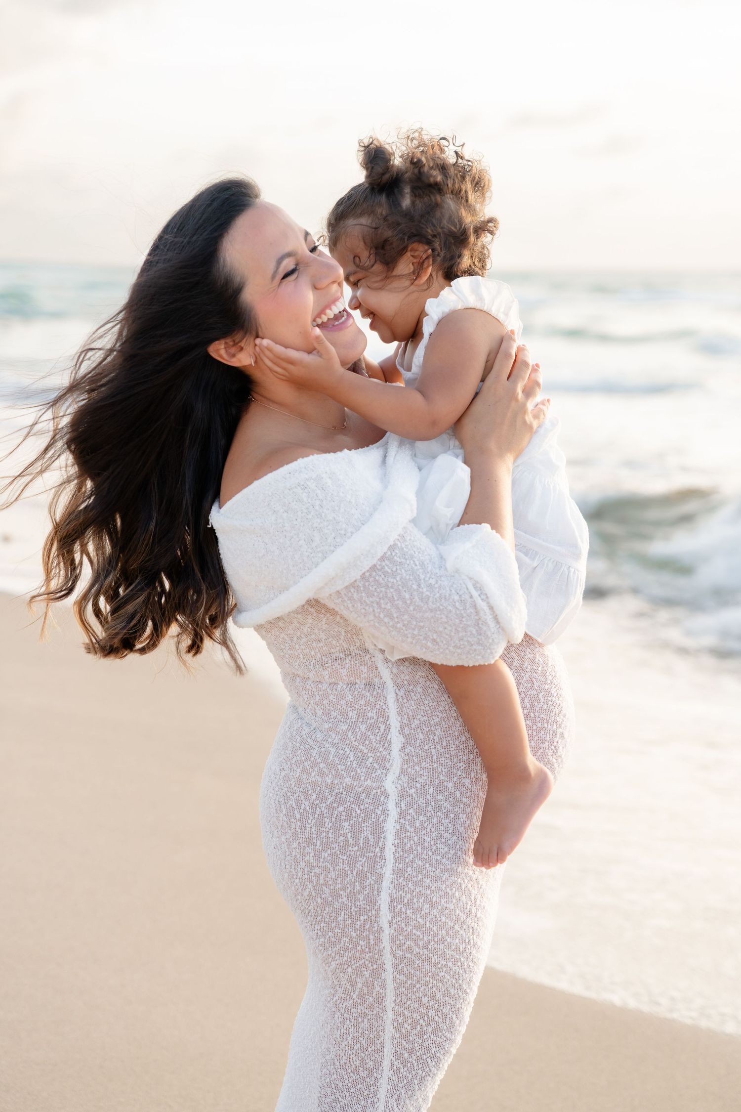 A joyful mother and child share a tender moment while wearing white dresses on a sunny beach.