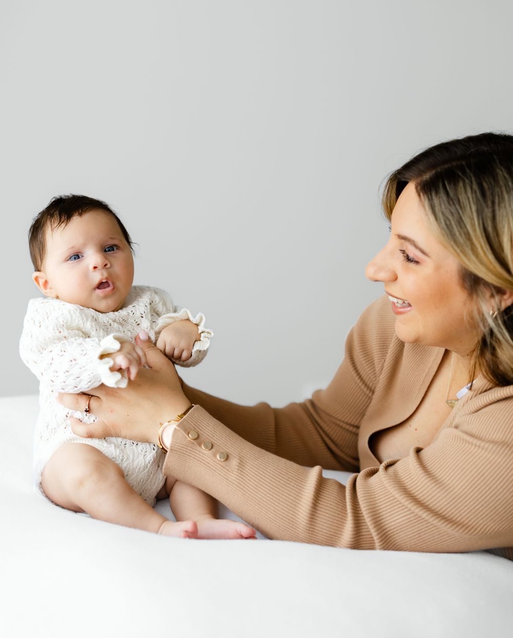 A parent and baby dressed in neutral colors share tender moments while lying on a light backdrop.