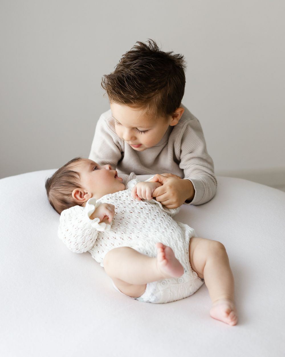 Two young children on white bed with little one wearing white knit onesie while older sibling gazes lovingly from above.