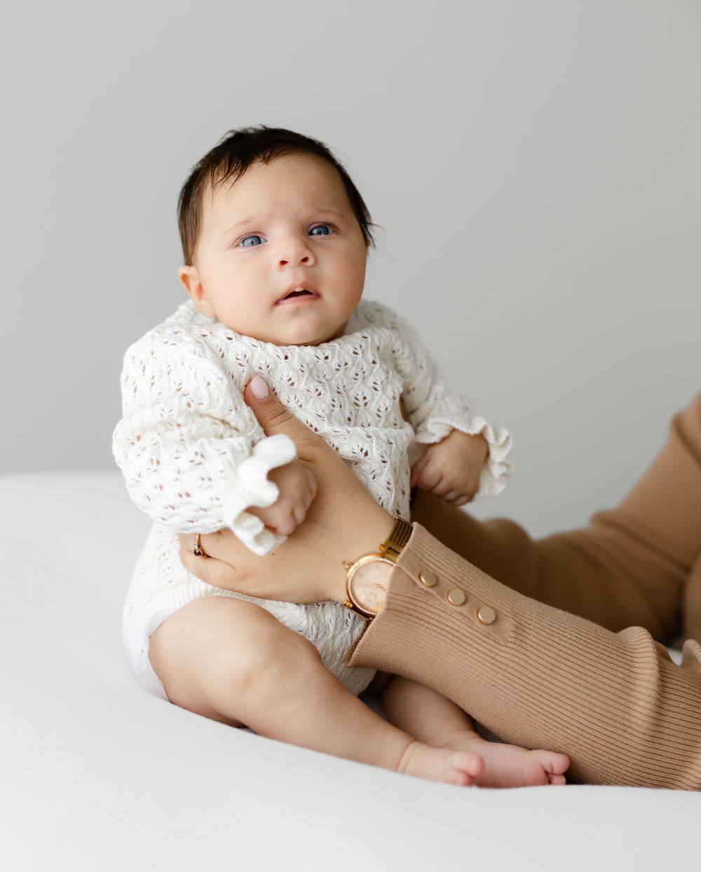 Baby in white knit sweater sits on white surface while being held by someone in tan sleeves.