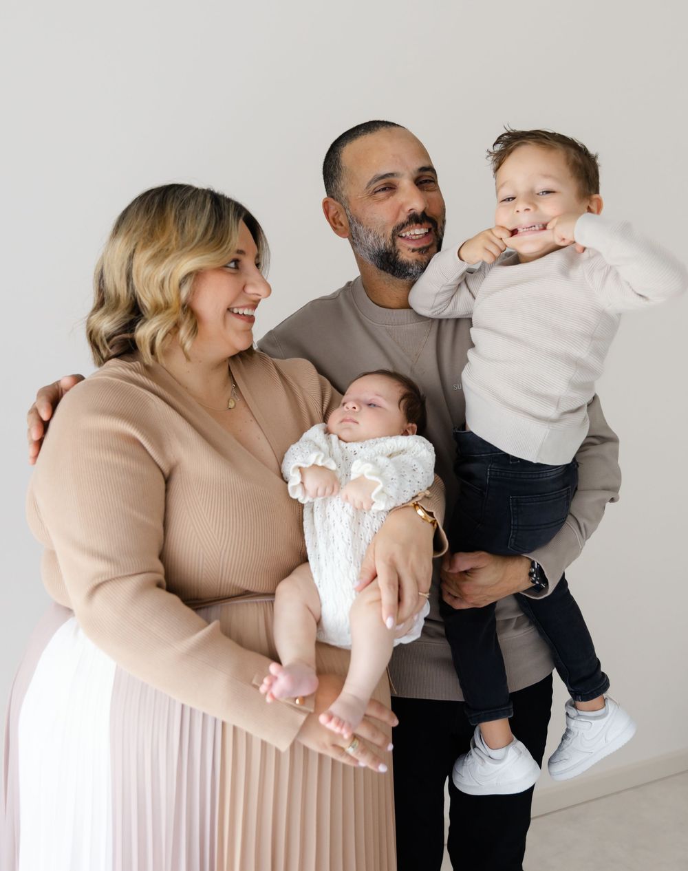 Family of four poses together against white wall in neutral-toned clothing.
