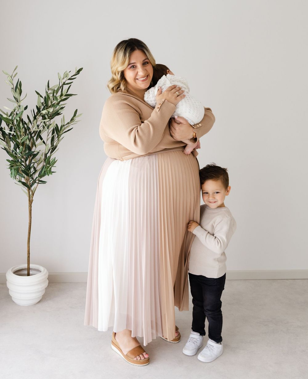 Mother in beige sweater and pleated skirt embraces baby while young child hugs her leg beside potted plant.