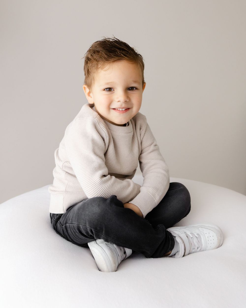 Young child in cream sweater and jeans sits cross-legged on white surface.