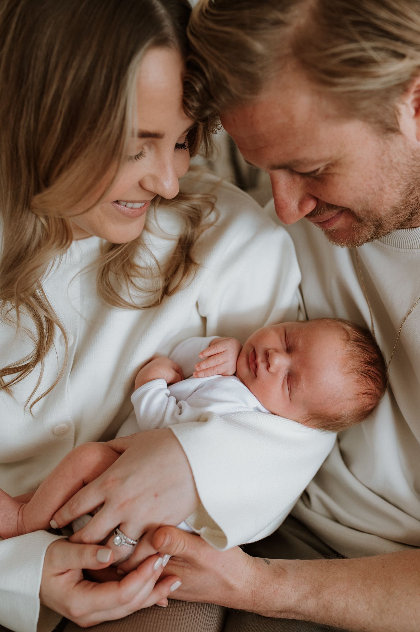 Intimate family portrait of parents in white sweaters lovingly gazing at their newborn baby.