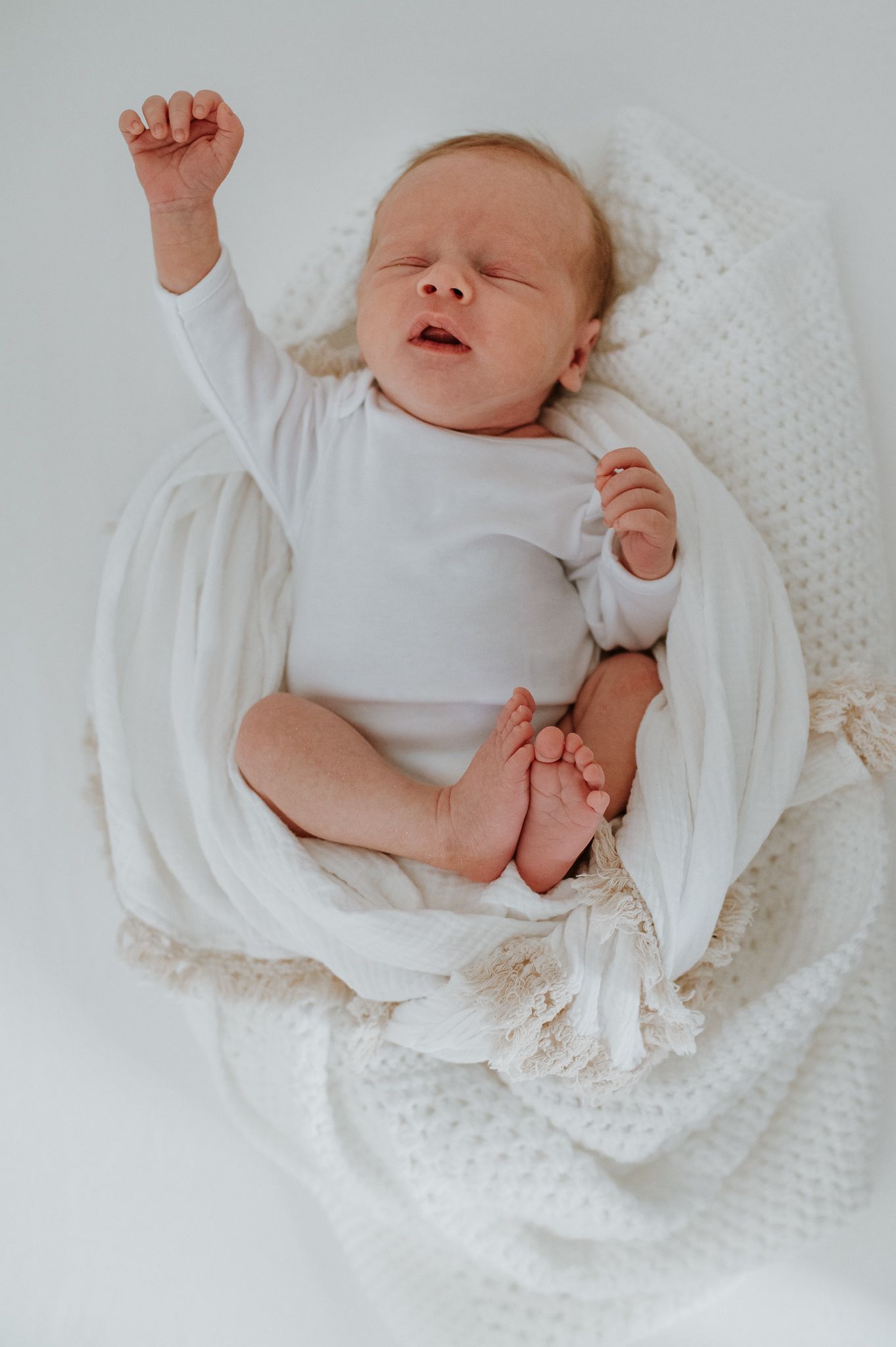 A newborn baby stretches and yawns while laying on soft white blankets during a sleepy moment.
