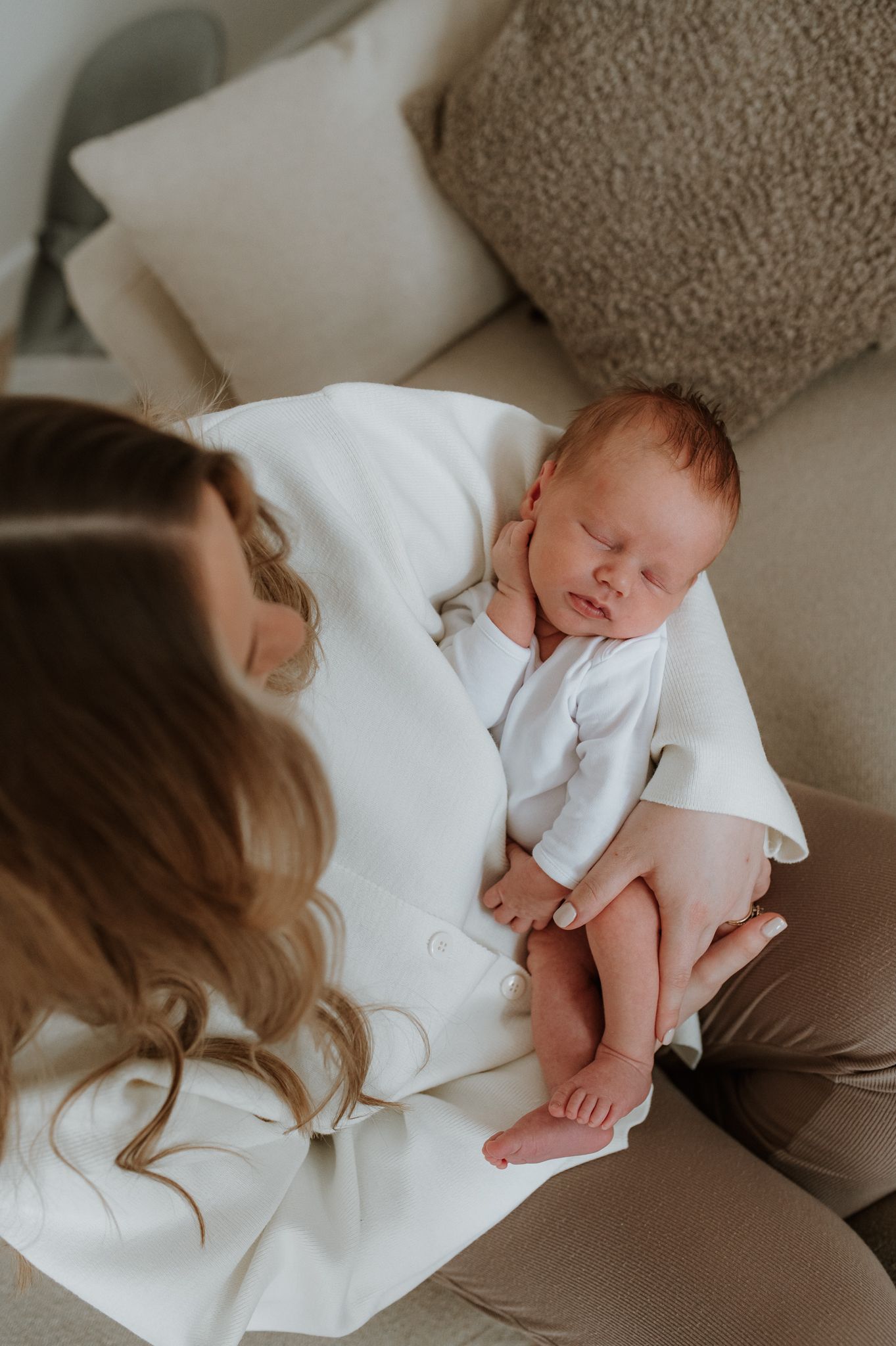 A newborn baby sleeps peacefully while wrapped in soft white blankets with a beige textured backdrop.