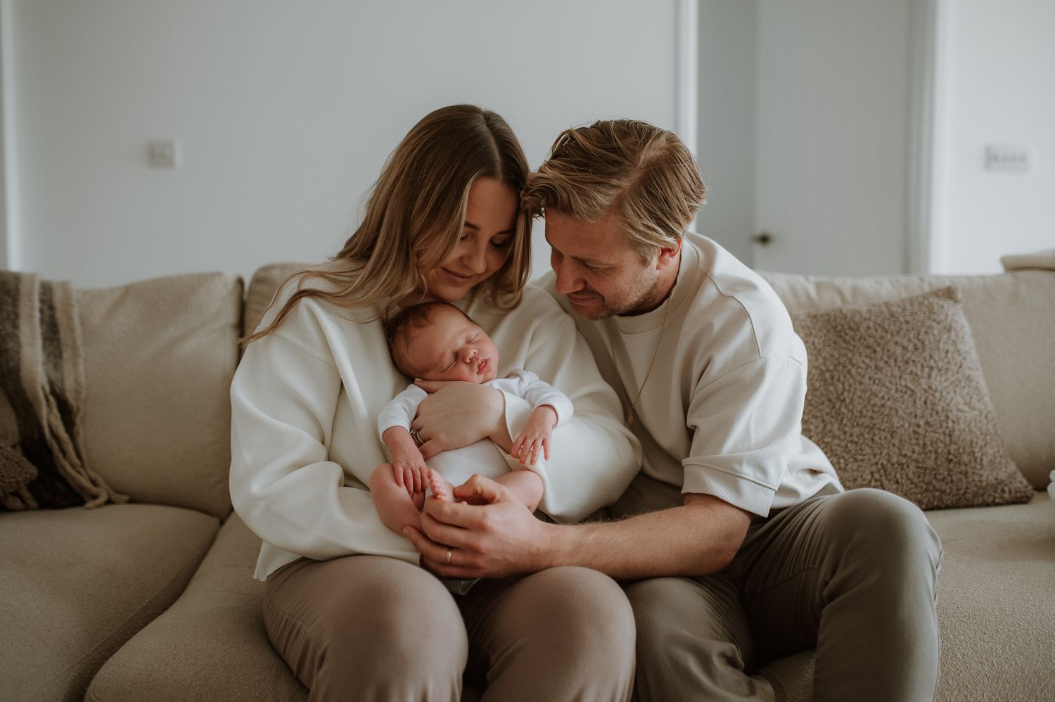Two people in white sweaters share an intimate moment with a baby while sitting together on a beige couch.
