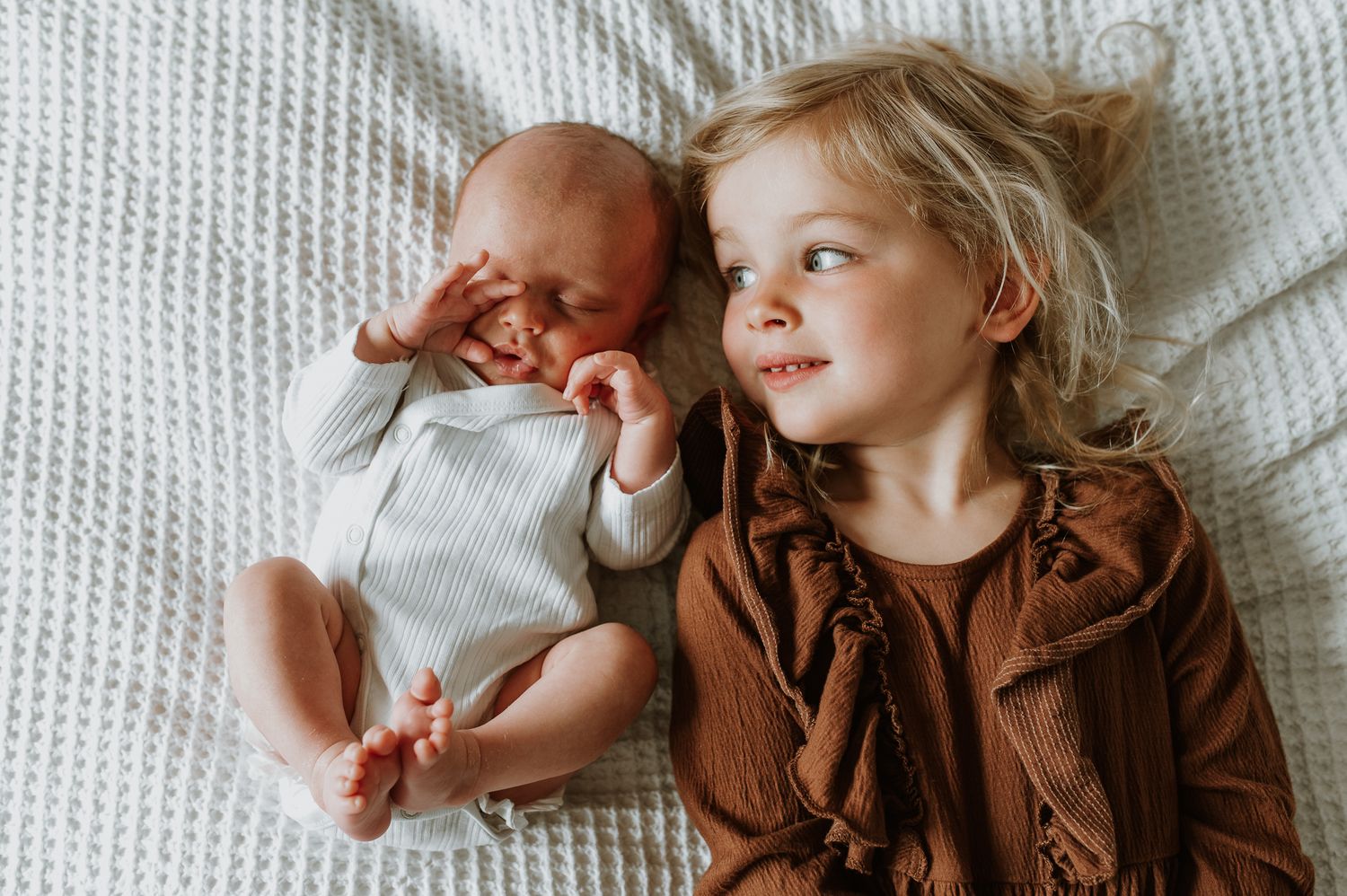 A newborn baby rests peacefully on white bedding in a natural light setting.