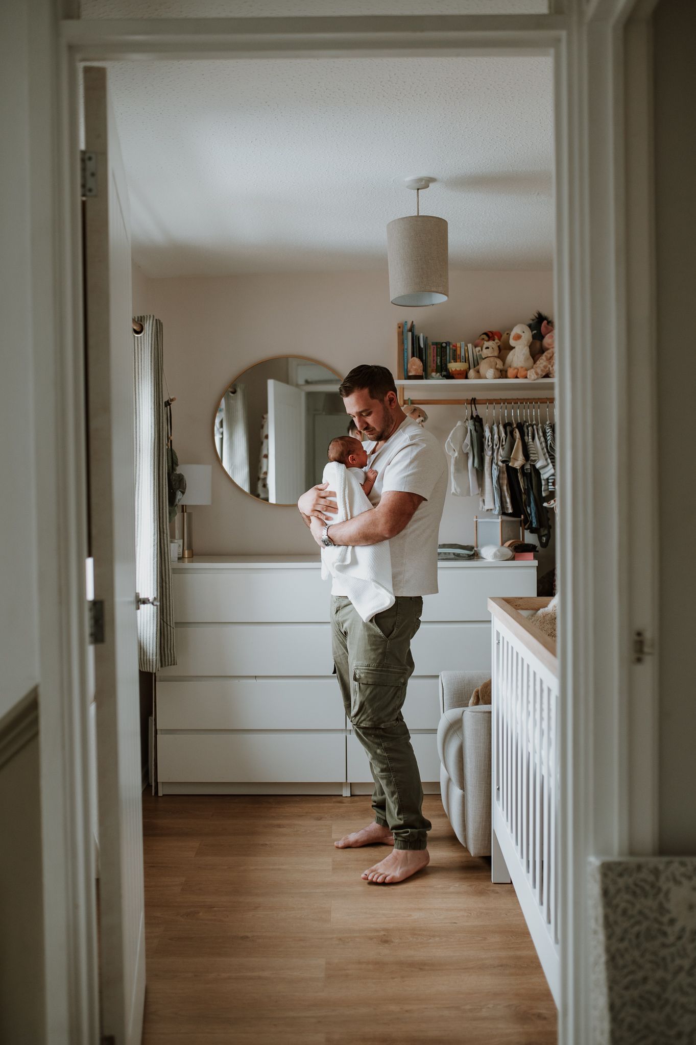 View through doorway into nursery with modern white furniture, hardwood floors and pendant light fixture.