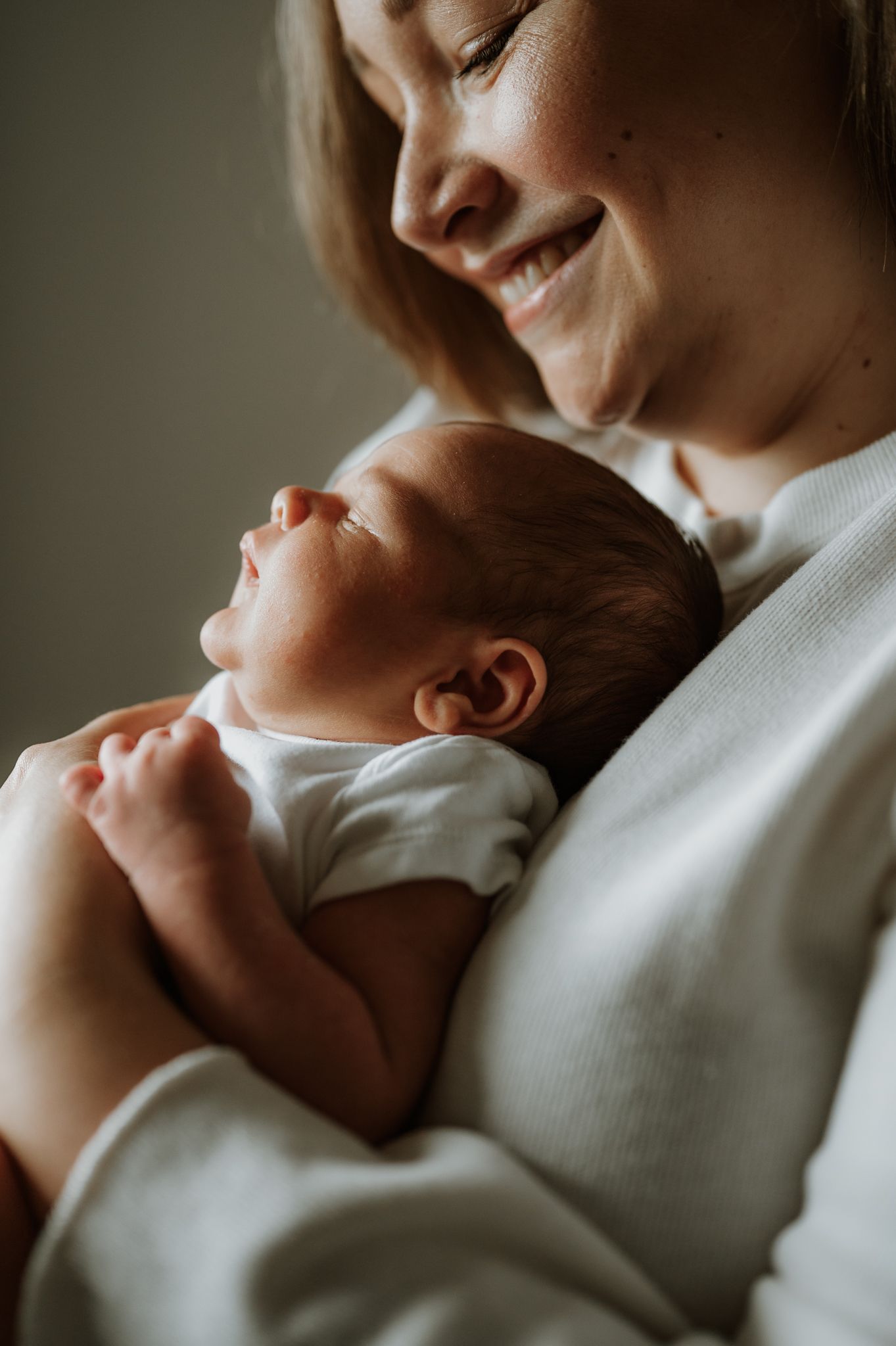 A tender moment between parent and newborn as they cuddle in soft natural light wearing white clothing.