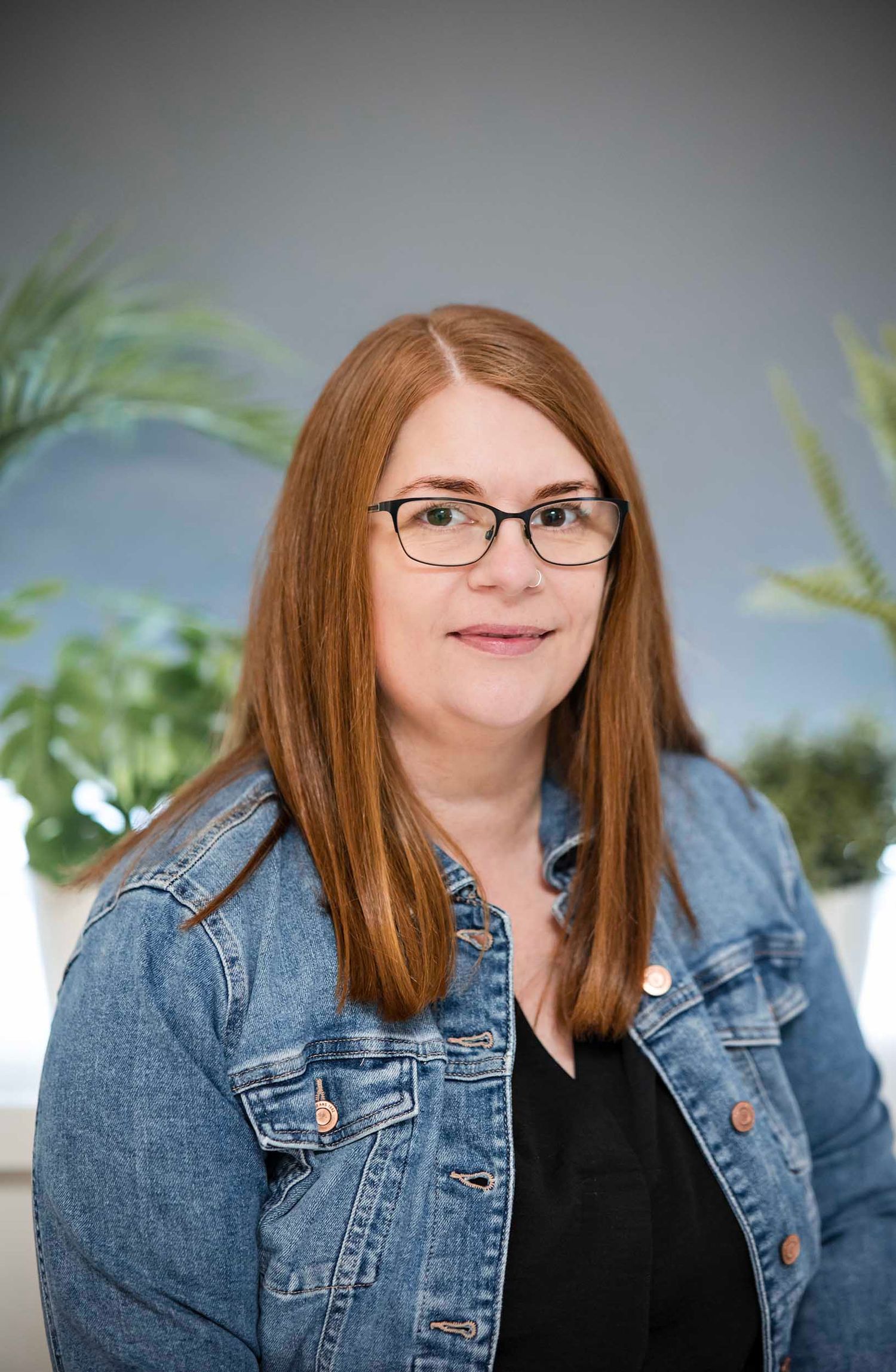 Professional indoor portrait showing someone with red hair wearing glasses and denim jacket against a light background. Tiny moments