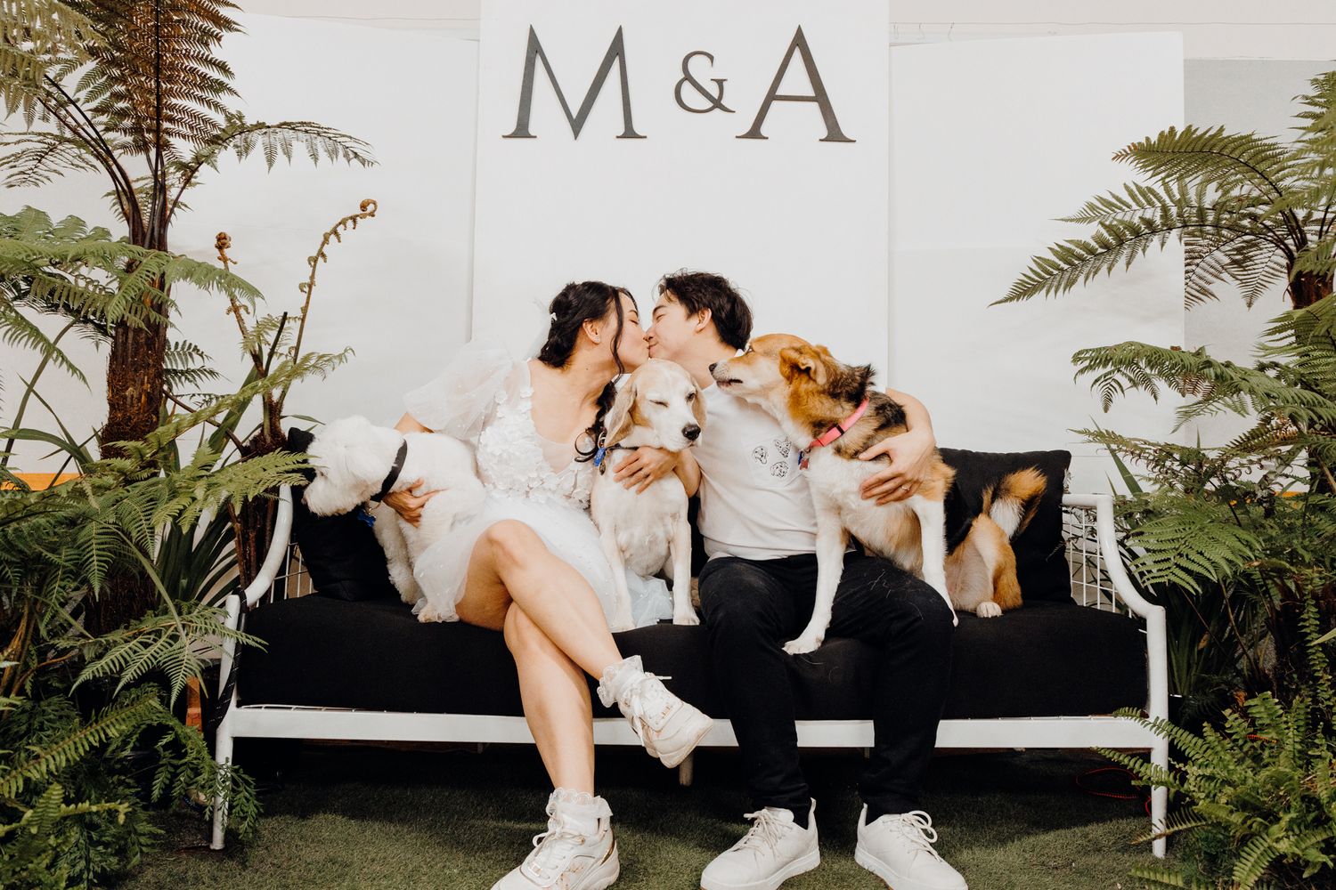 Couple sits on white bench with dogs surrounded by tropical plants and M&A signage.
