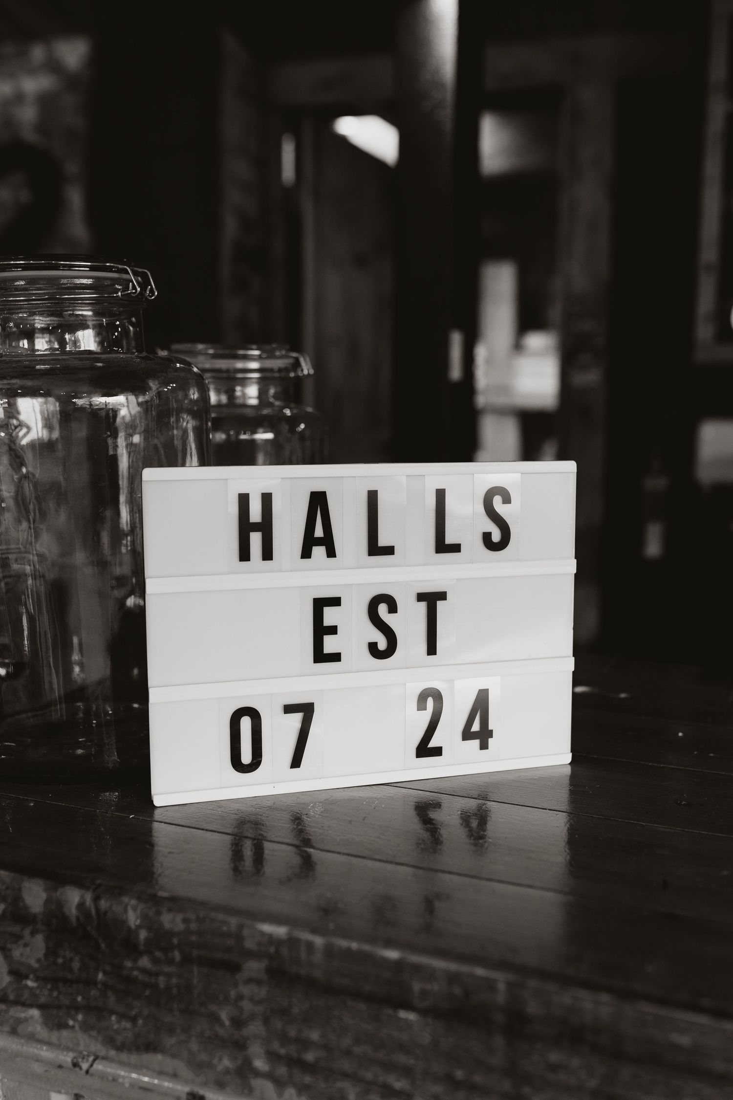 A black and white photo of a lightbox sign reading 'HALLS EST 07 24' with glass jars visible in the background.