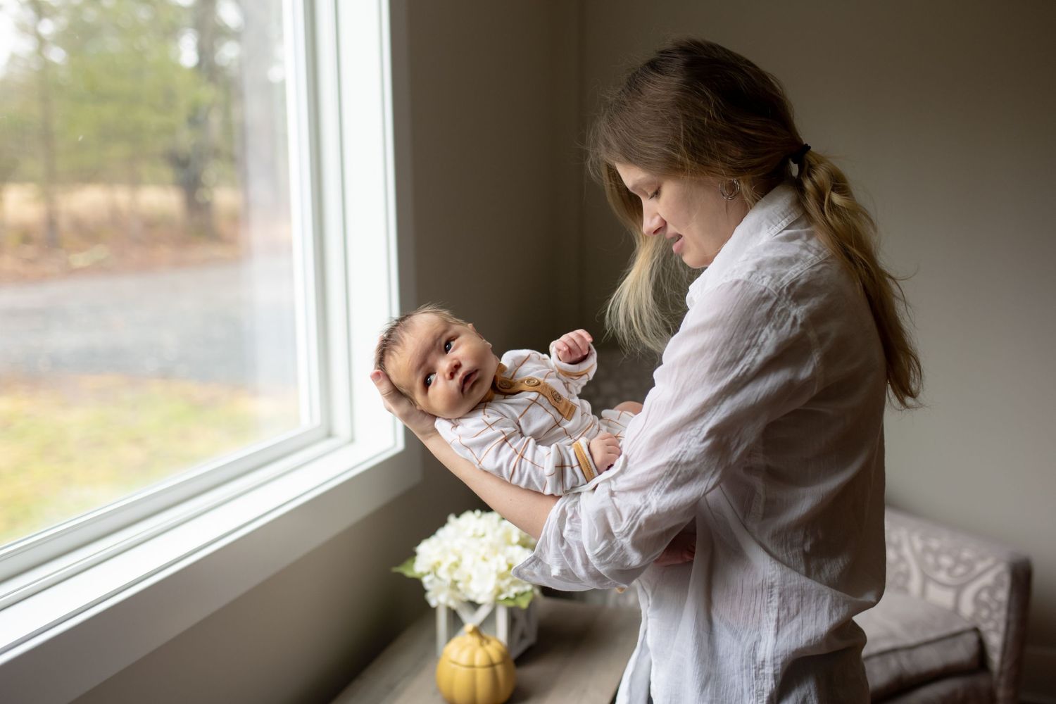 A parent holds a newborn baby while standing by a window with soft natural light streaming in and flowers on the windowsill.