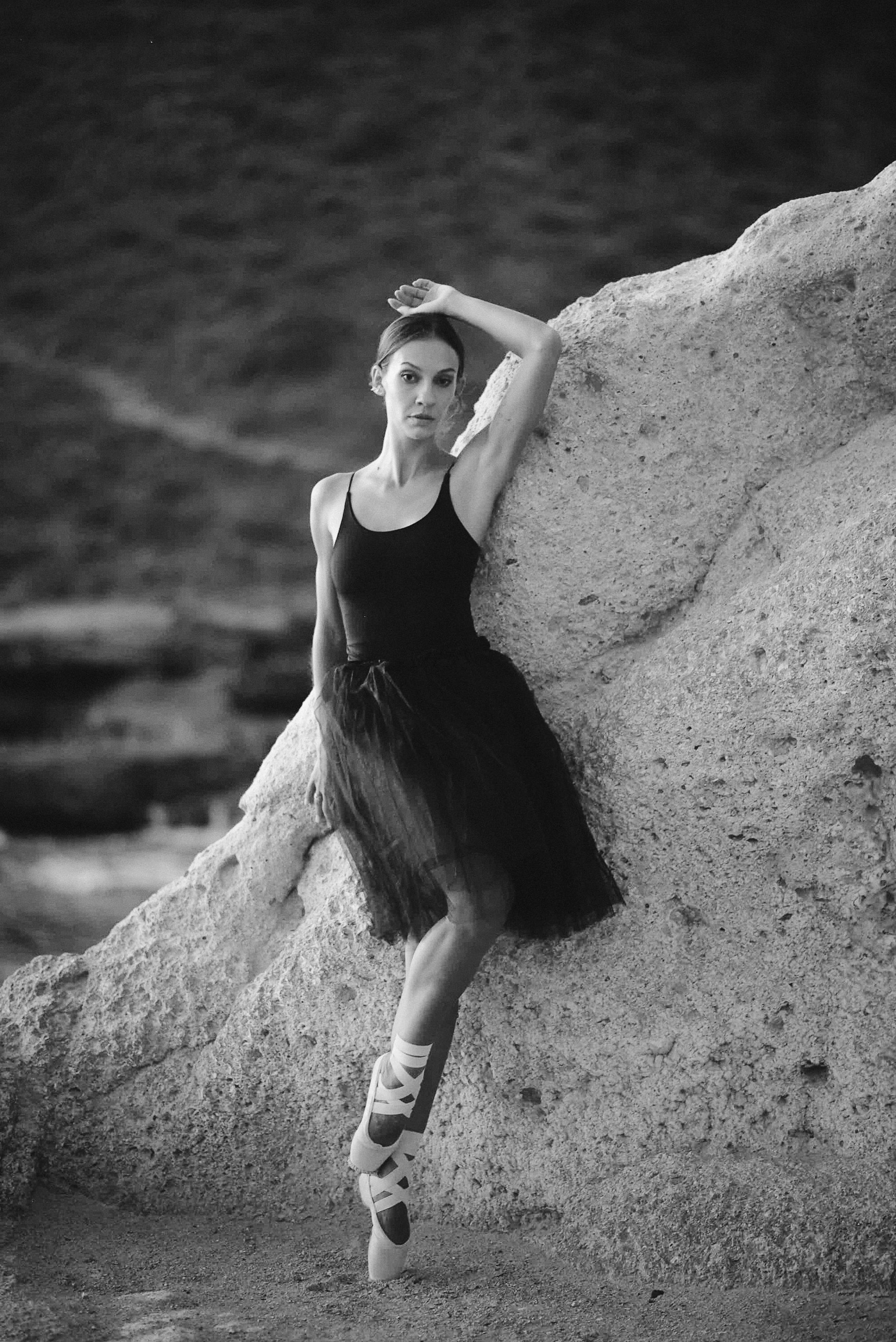 A ballet dancer in black tutu performs en pointe against a rocky coastal backdrop. Tenerife Wedding Photographer in La Caleta de Adeje.