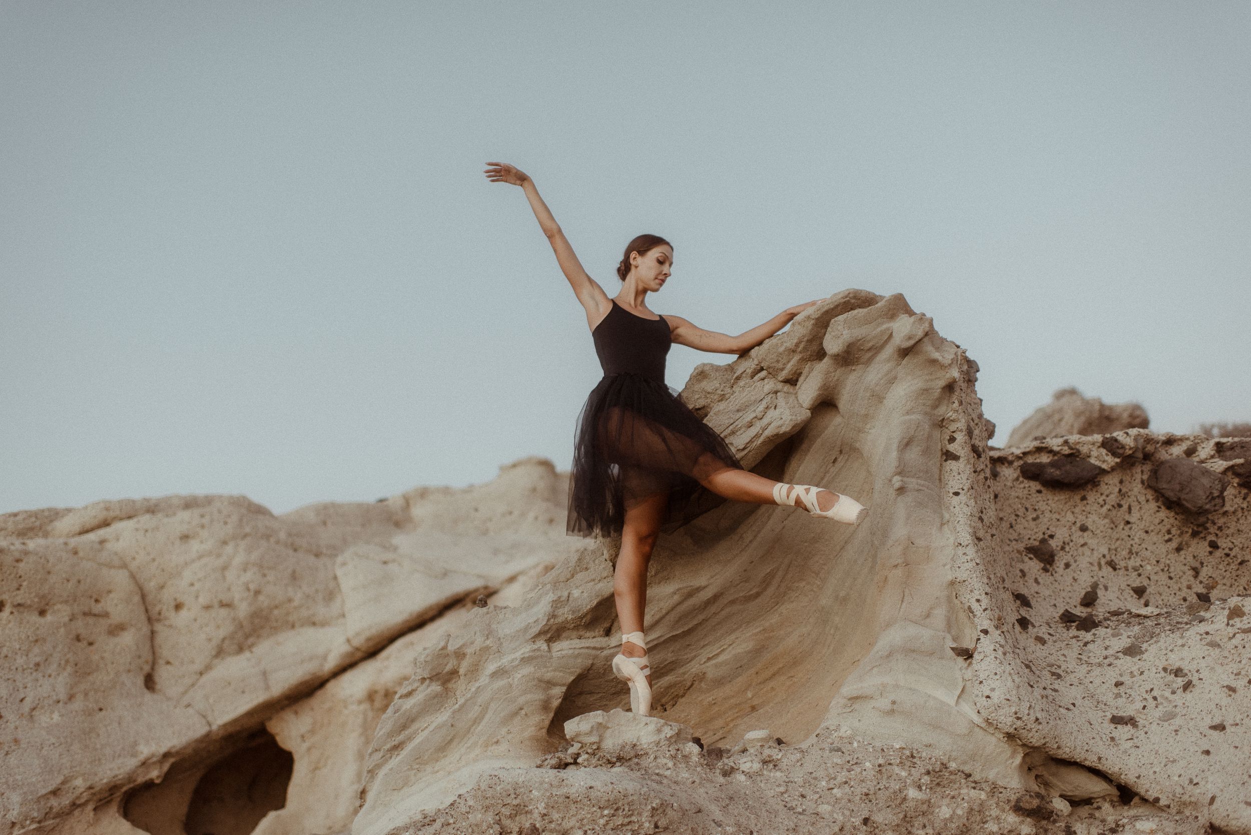 A ballet dancer in a black dress poses gracefully on rocky terrain with flowing fabric in the wind. Tenerife Wedding Photographer