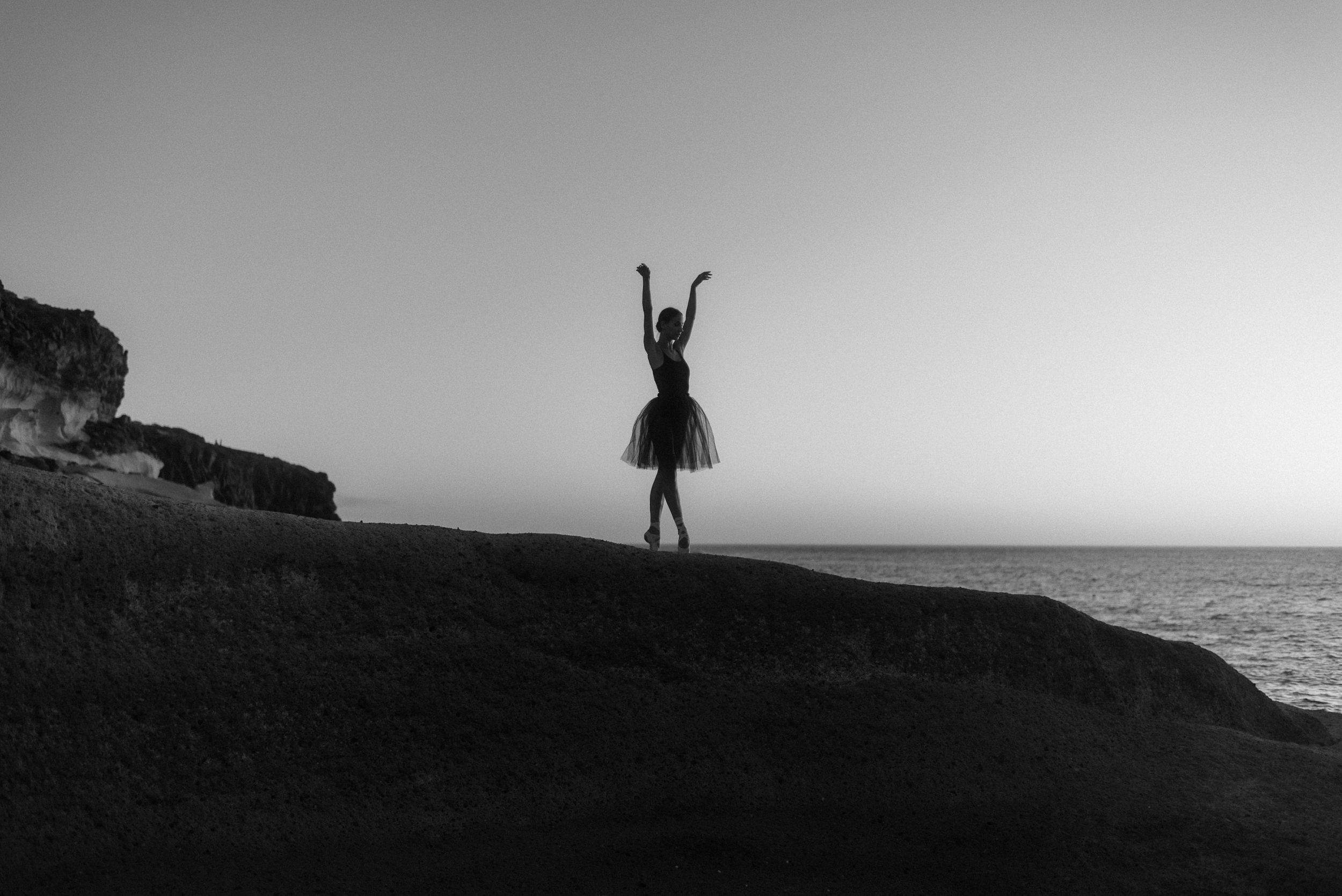 A silhouetted ballet dancer poses gracefully against a sunset sky on rocky coastal cliffs. Tenerife Wedding Photographer in La Caleta