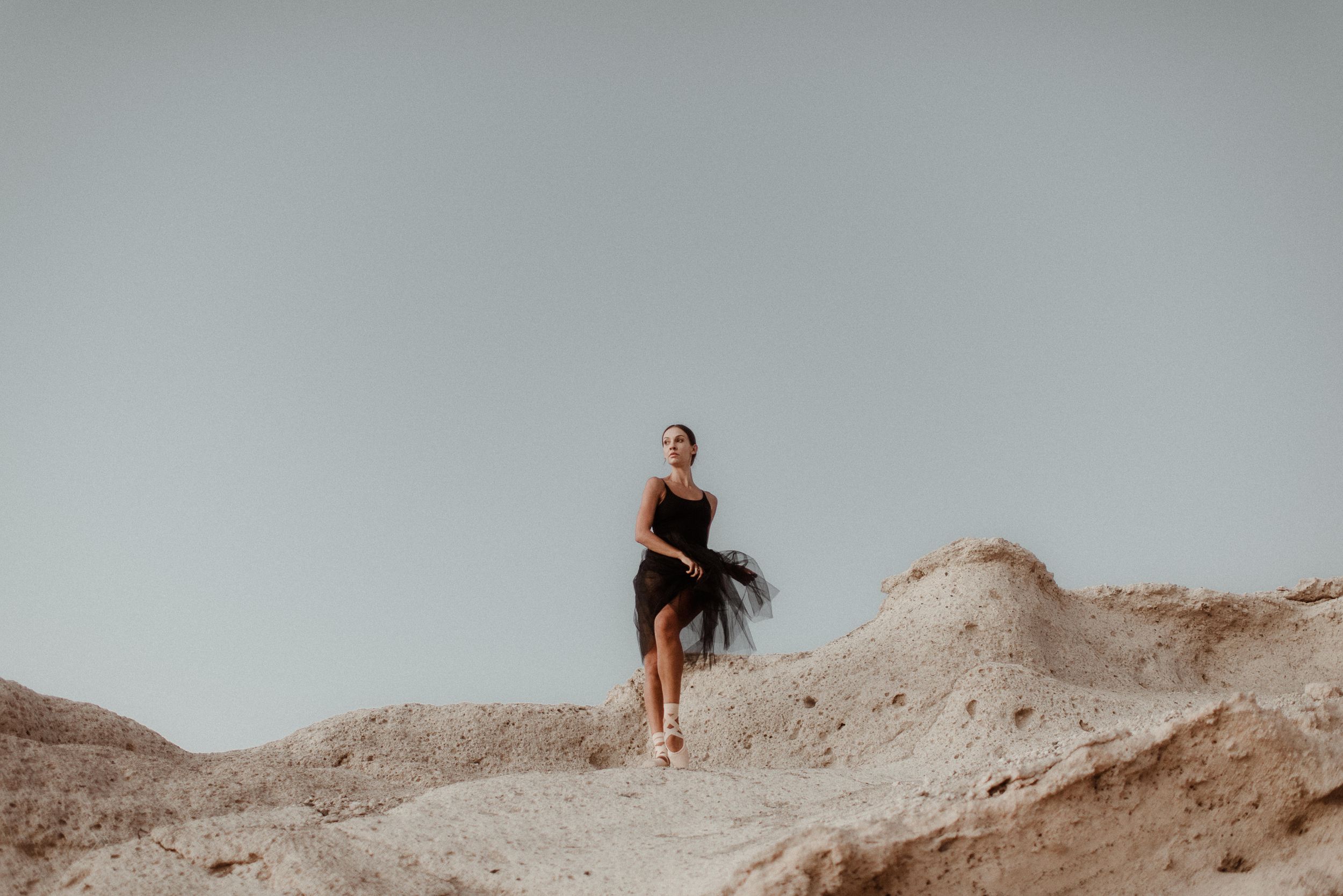 A dancer in a black tutu poses elegantly on sandy dunes against a light background. Tenerife Wedding Photographer in La Caleta de Adeje.