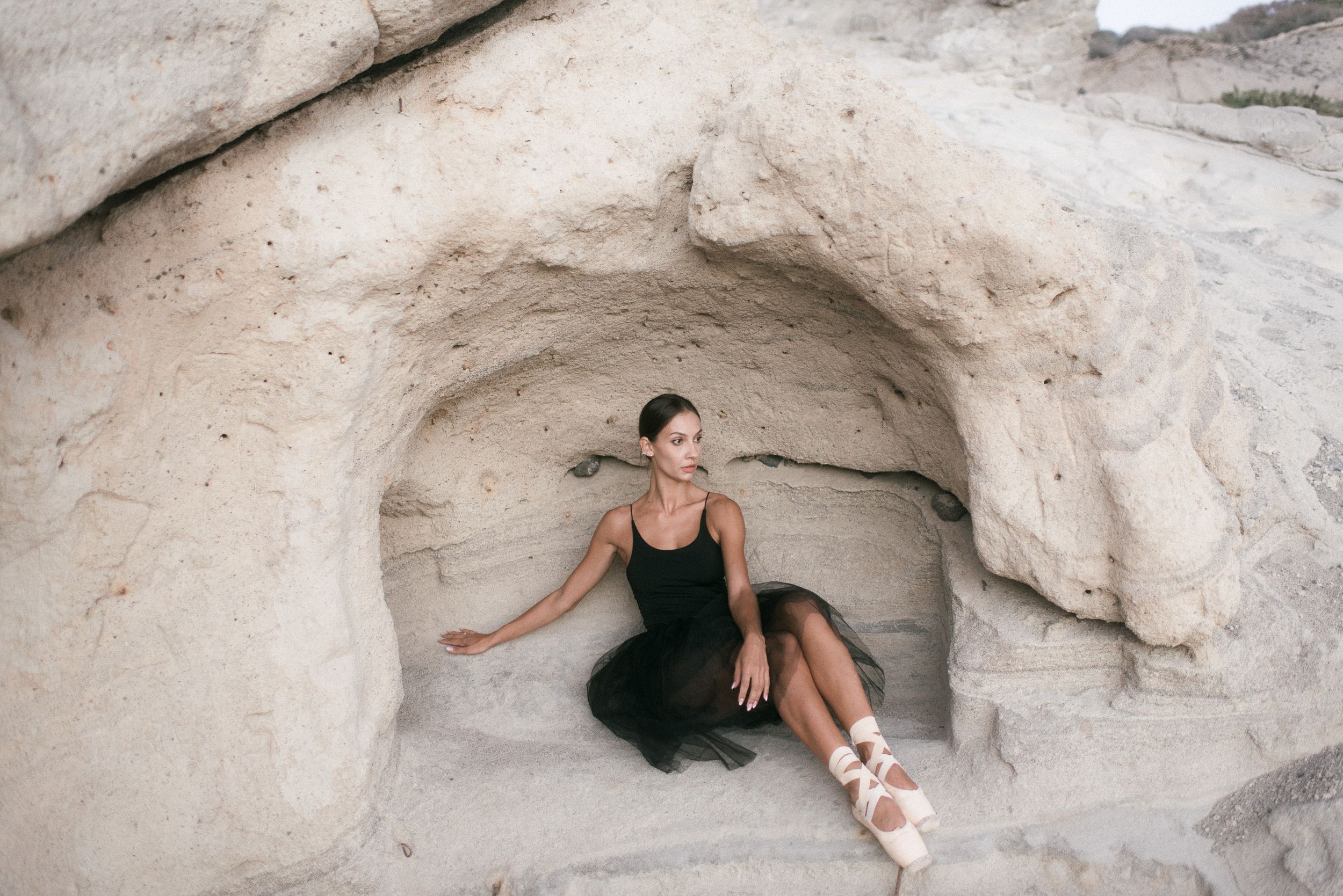 Ballerina in black dress sits elegantly in white stone cave formation with pointe shoes. Tenerife Wedding Photographer in La Caleta de Adeje