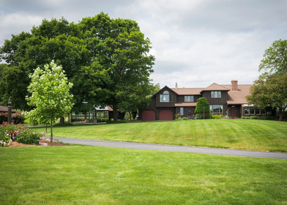 Large rustic home with brown siding and dormers sits on a manicured lawn with mature trees and curved driveway.