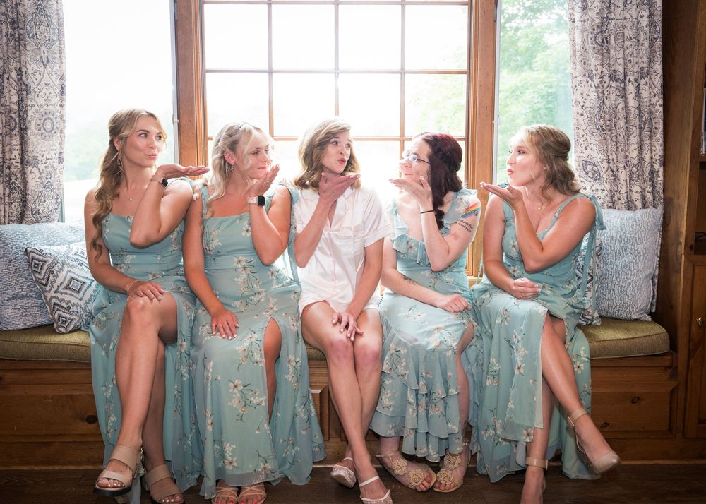 Bridesmaids in matching blue dresses share laughter while sitting on window bench.
