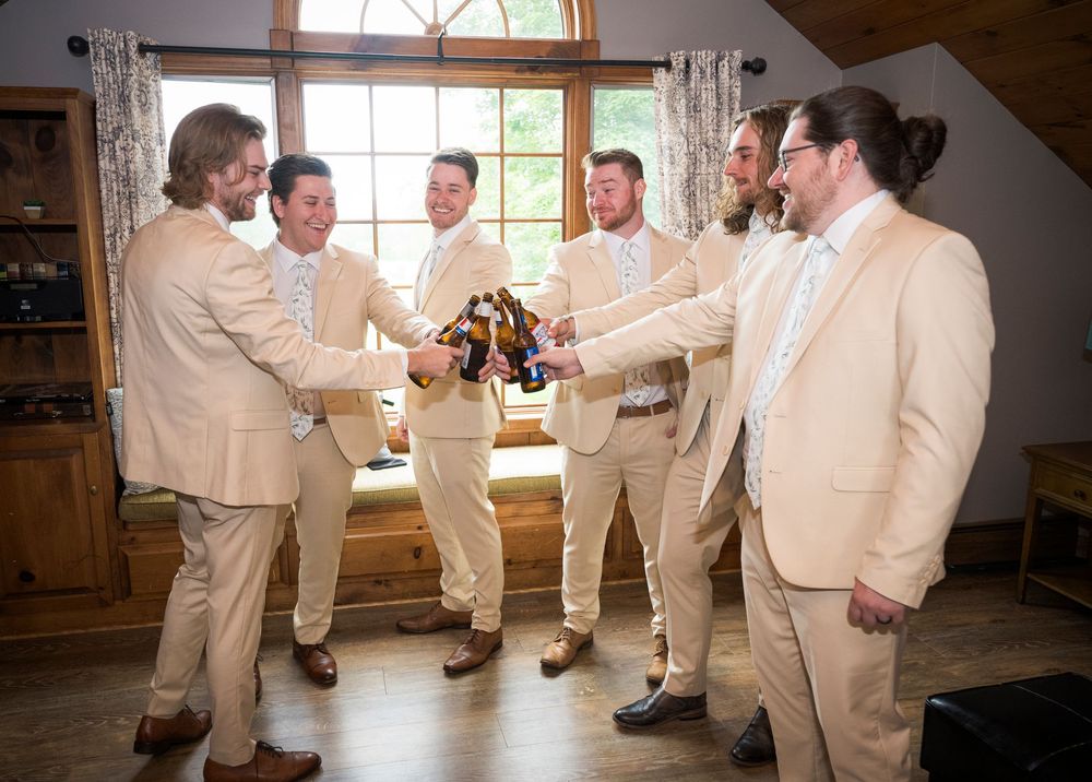 Groomsmen in beige suits toast with beer bottles in a rustic cabin setting.