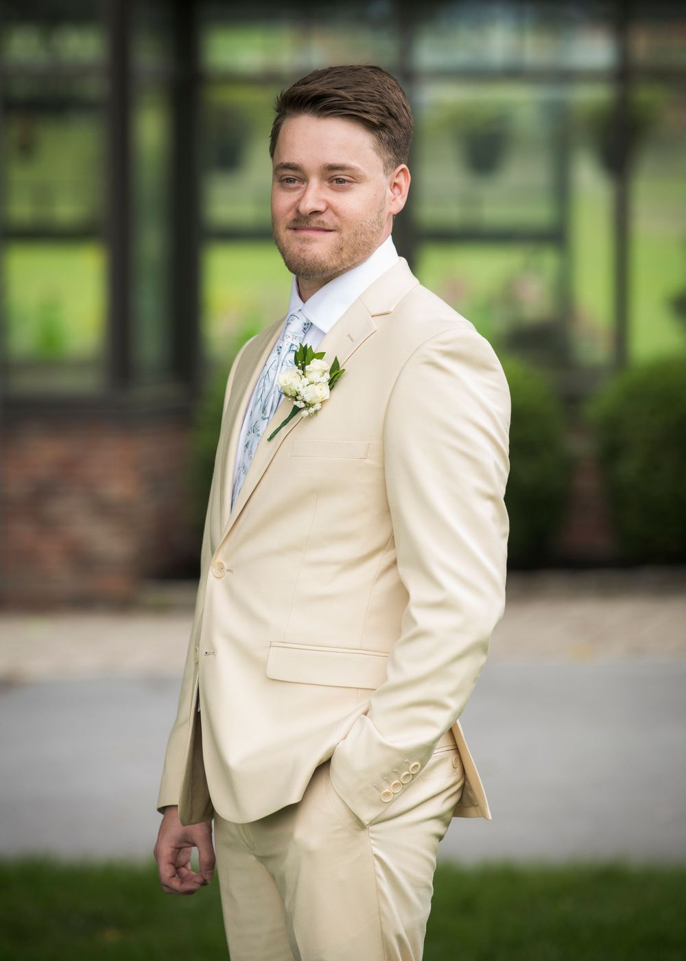 Groom in a cream colored suit with white boutonniere stands outdoors at formal event.