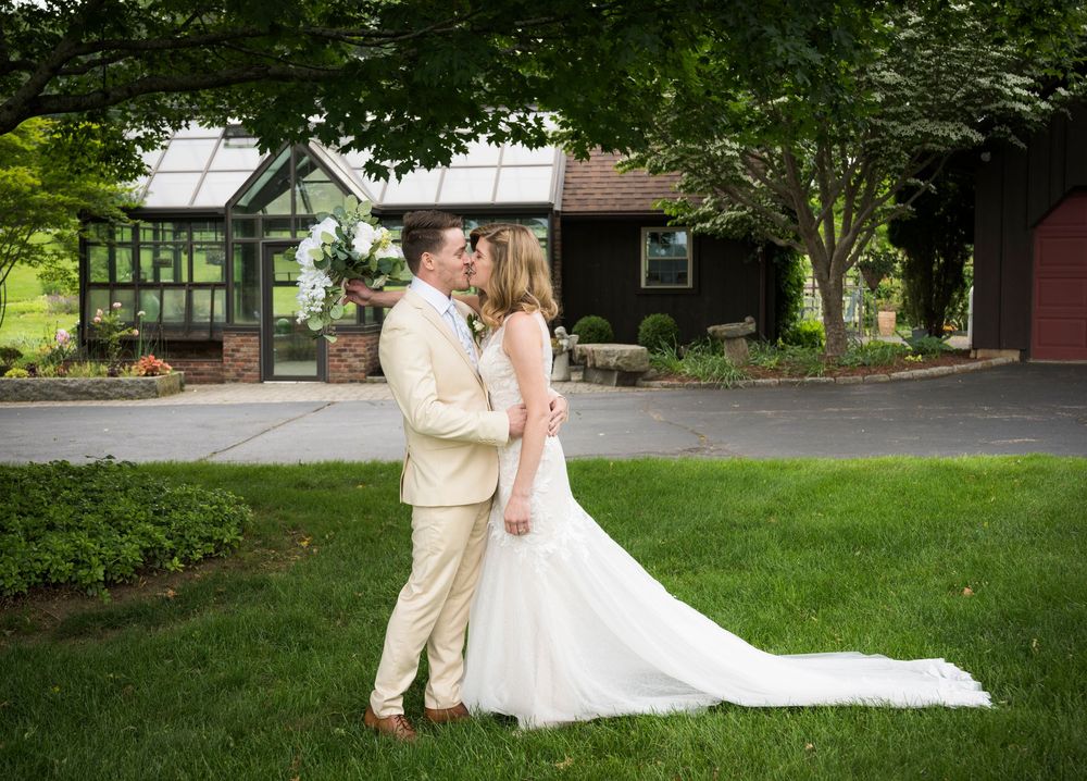 Wedding couple embraces on green lawn with rustic buildings in background.