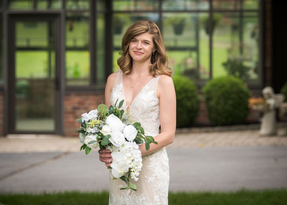 A bride in a lace v-neck wedding gown holds an elegant white floral bouquet in front of a window-filled venue.