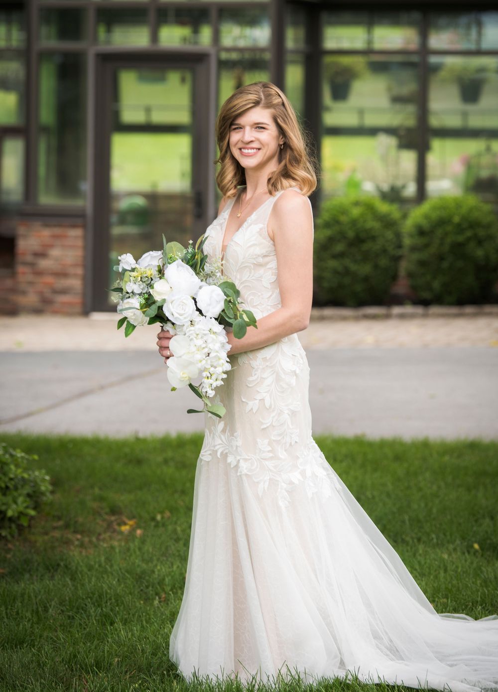 A bride in a fitted lace wedding gown holds a white floral bouquet against a garden backdrop.