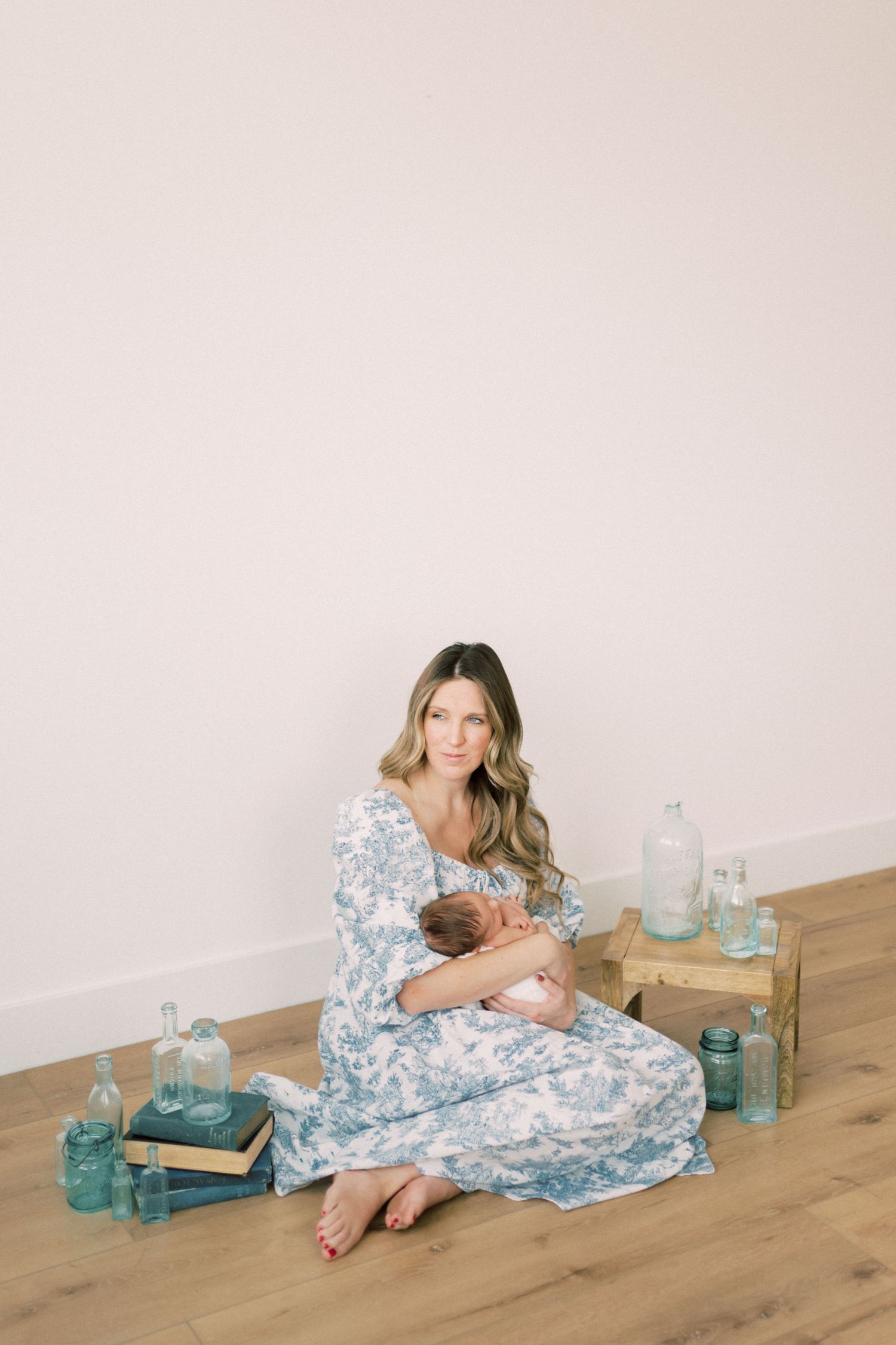 Person in a blue and white floral dress sits on wooden floor with vintage glass jars and decorative elements.