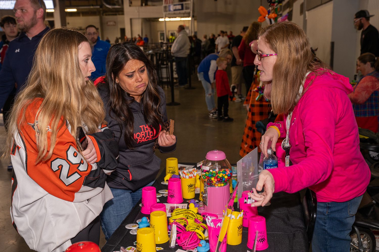 People gathered at tables with yellow and pink toys and merchandise on display at a community event.