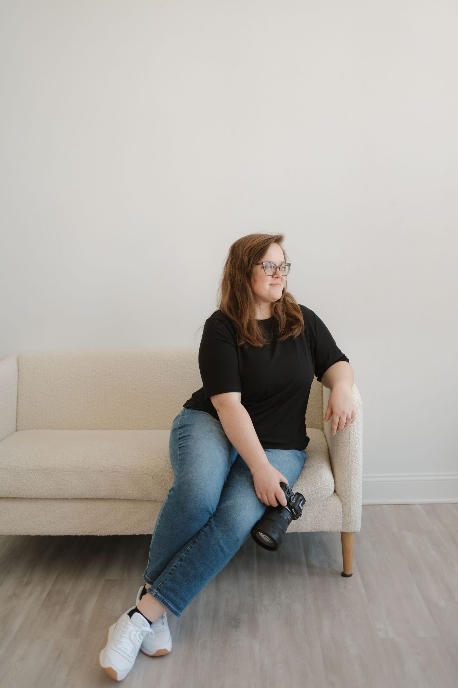 Person in casual attire with glasses sits casually on a light beige couch against a white wall.