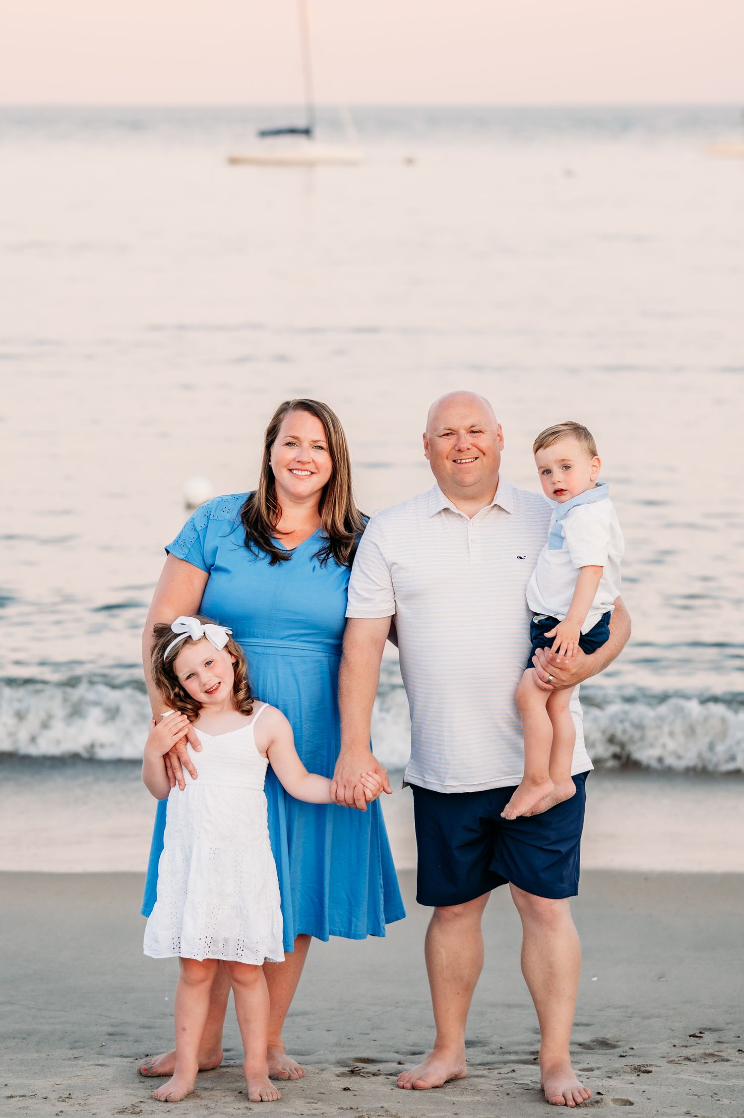 Family portrait on a beach at sunset with ocean waves and sailboat in background.