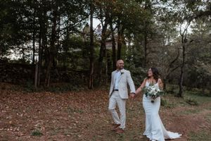 A couple walks hand in hand through a wooded area in elegant wedding attire.