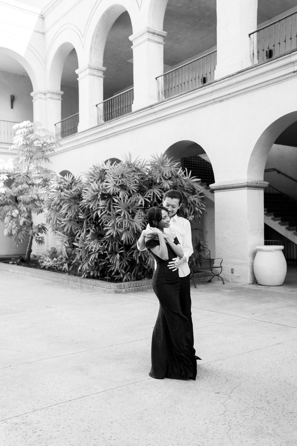Black and white photo of a couple in formal attire standing under arched colonnade with tropical plants in background.