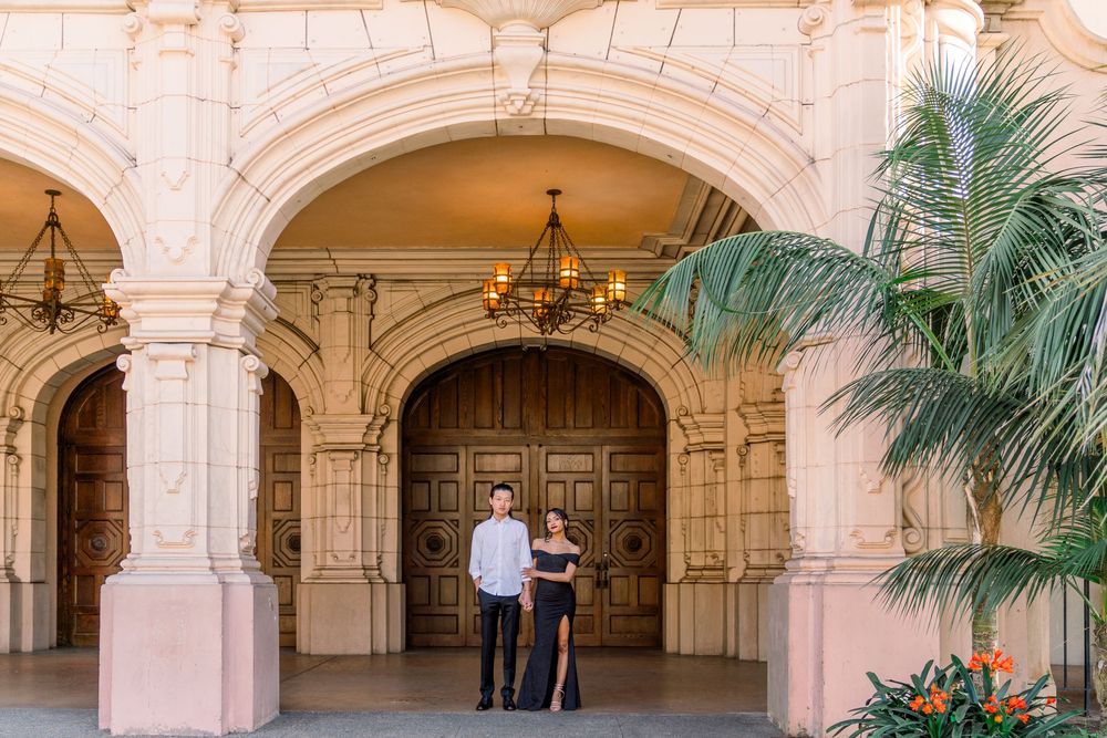Two people stand in front of an ornate archway with decorative columns and a palm tree at Balboa Park in San Diego.