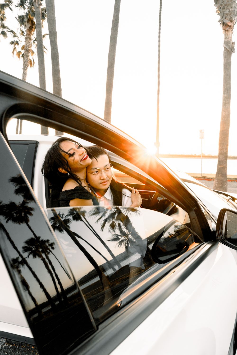 Palm trees reflect on white car window while passengers enjoy a golden sunset drive near beach.