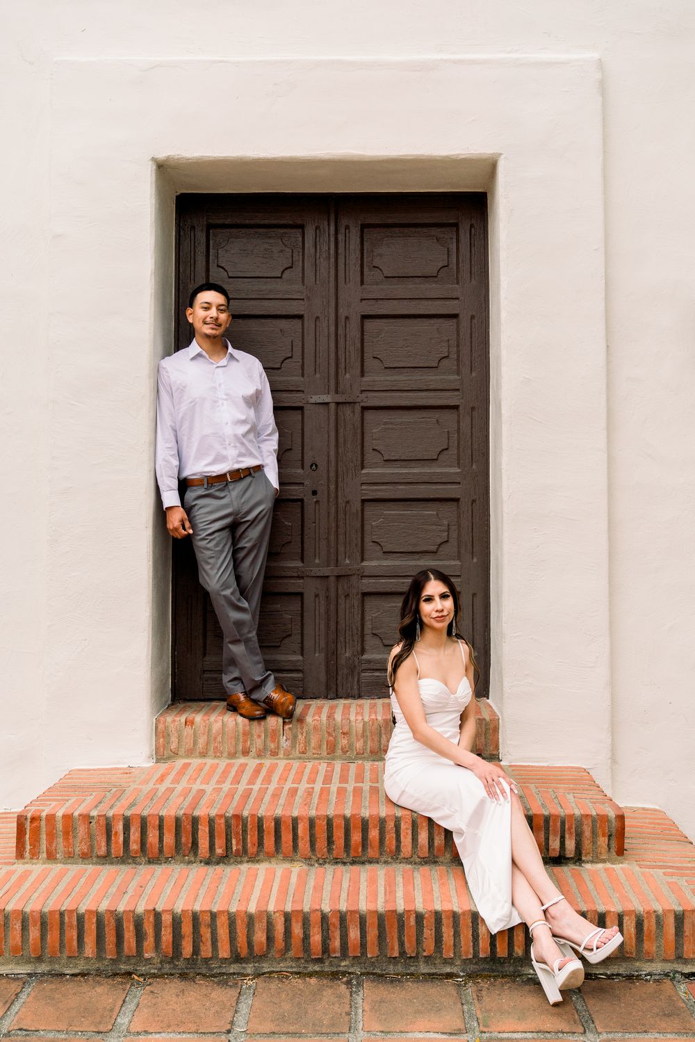 A couple in formal attire poses on brick steps in front of a large dark wooden door against a white building.